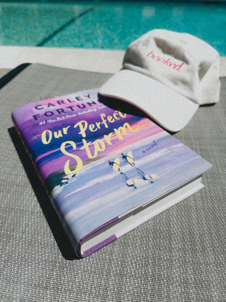 A close-up of the book 'Our Perfect Storm' by Carley Fortune, resting on a textured surface next to a beige cap with the word 'booked' embroidered in red. A swimming pool is visible in the blurred background.