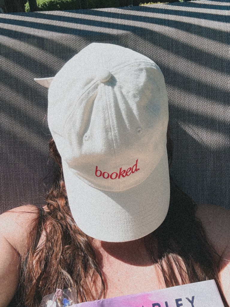 A person with long hair wearing a white baseball cap with the word 'booked.' printed in red, sitting outdoors in a sunny setting.